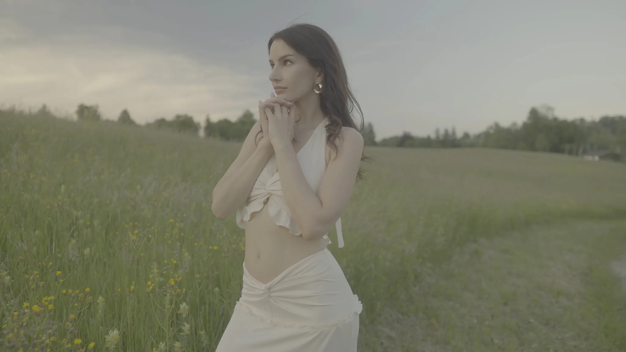 An ungraded frame: a woman standing in a summer meadow under a flat grey sky, washed out and low in contrast.